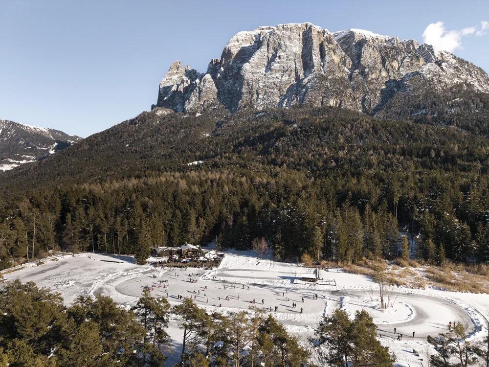 Menschen beim Schlittschuhlaufen auf einer Eisbahn vor bewaldetem Bergpanorama