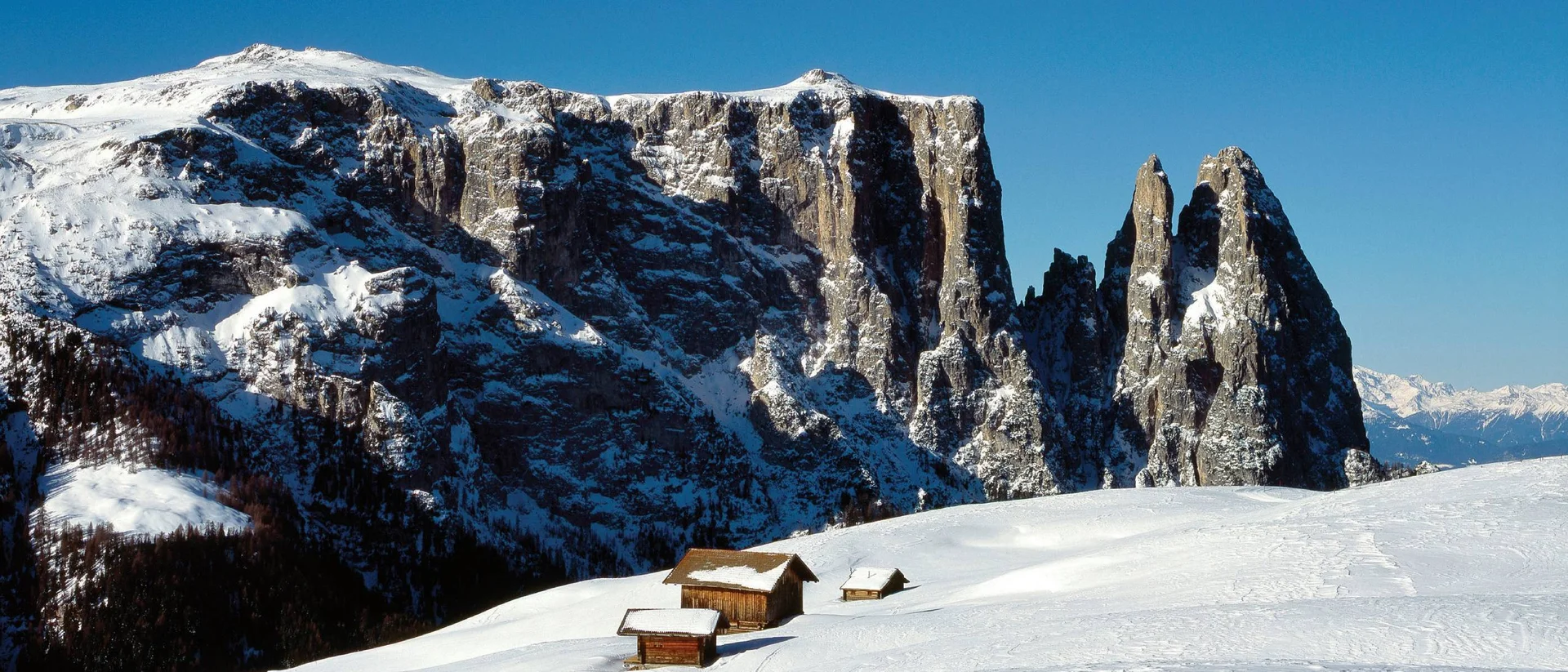 Schneebedeckte Alm mit Holzhütten vor felsigen Berggipfeln unter blauem Himmel