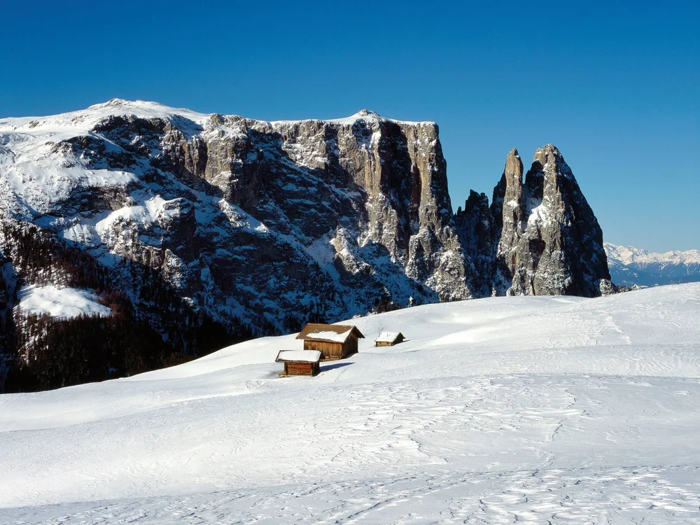 Schneebedeckte Alm mit Holzhütten vor felsigen Berggipfeln unter blauem Himmel