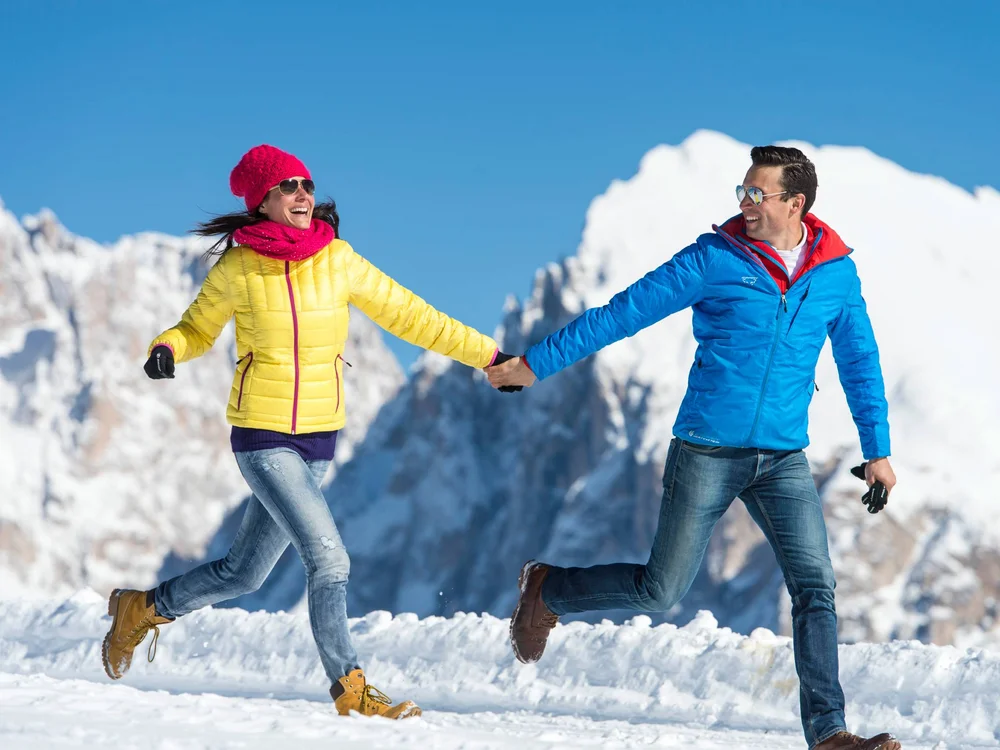 Paar in bunten Jacken läuft lachend im Schnee vor Bergkulisse