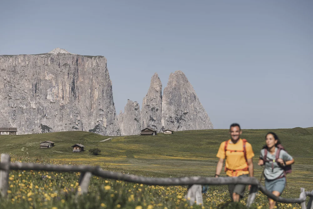 Wanderer auf grünem Hügel mit Bergpanorama und Hütten im Hintergrund