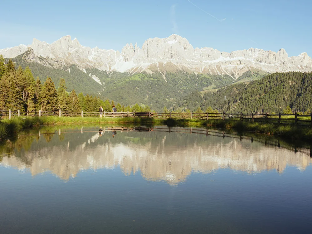 Bergkette mit Spiegelung im See und zwei Personen am Zaun