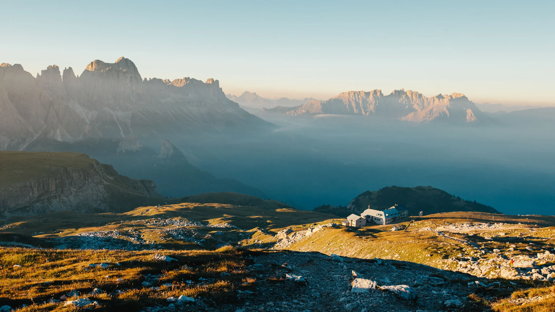 Berghütte auf sonnigem Hang mit Dolomiten im Hintergrund bei klarem Himmel