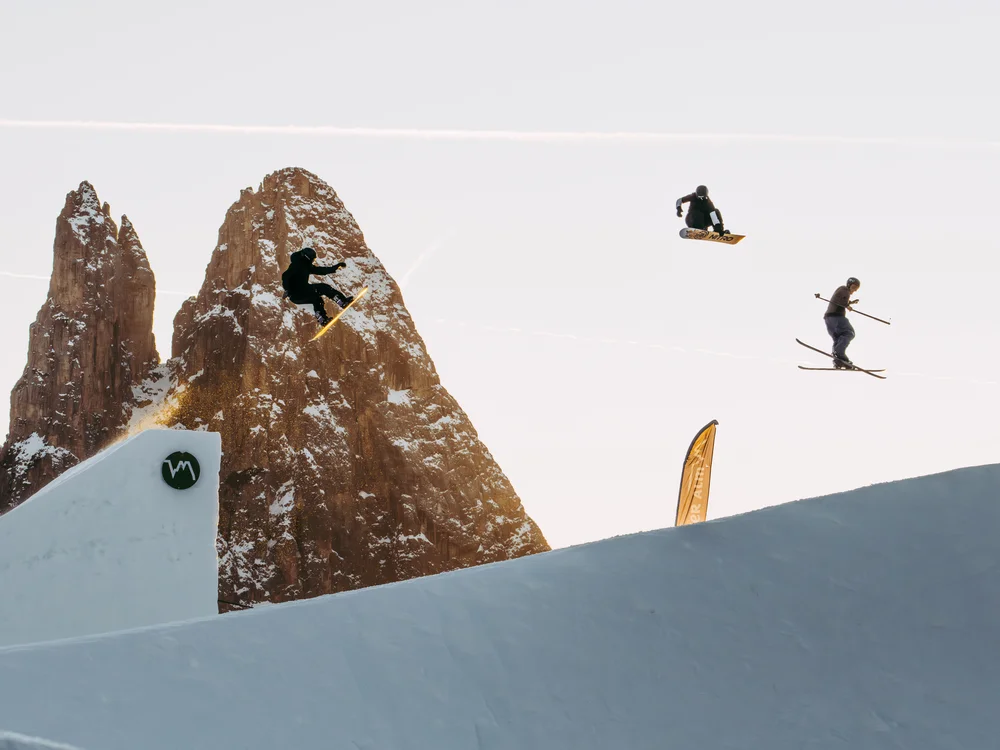 Drei Wintersportler springen mit Ski und Snowboard vor Berggipfeln