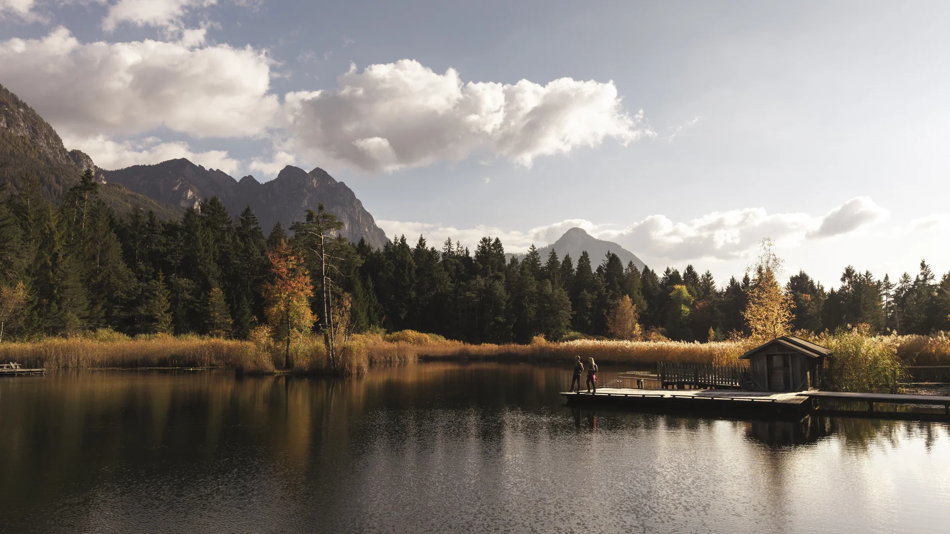 Bergsee mit Wald, Herbstbäumen, Steg und Hütte unter bewölktem Himmel