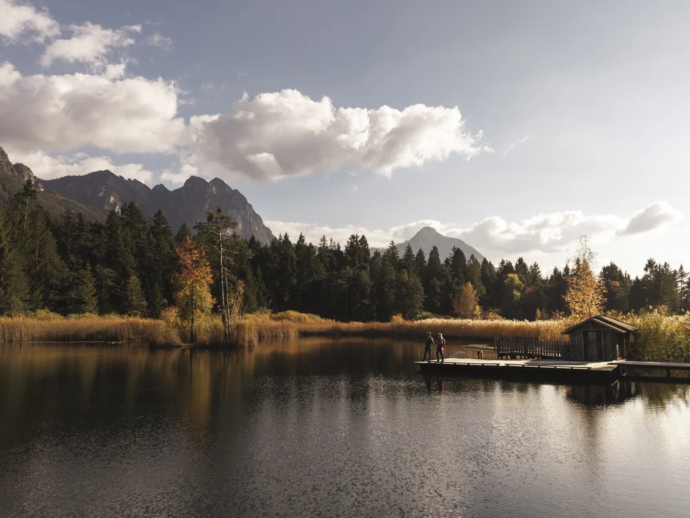 Bergsee mit Wald, Herbstbäumen, Steg und Hütte unter bewölktem Himmel