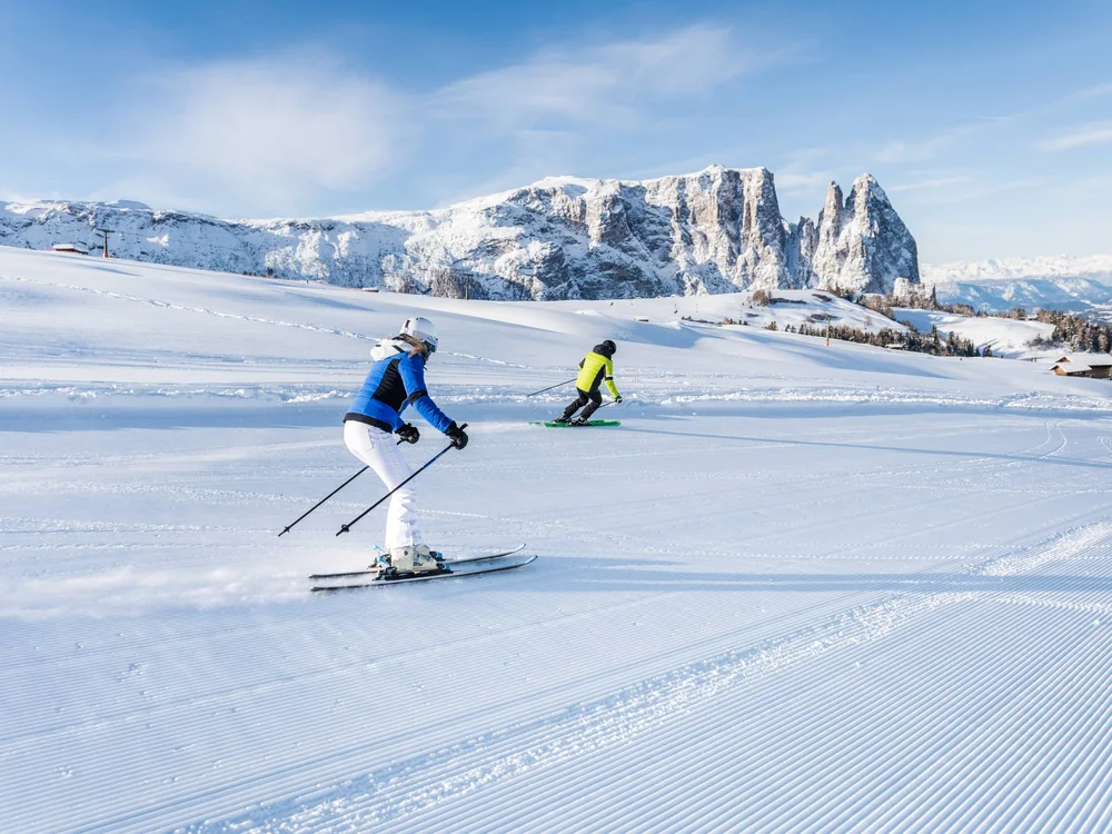 Zwei Skifahrer fahren auf einer präparierten Skipiste mit schneebedeckten Bergen im Hintergrund