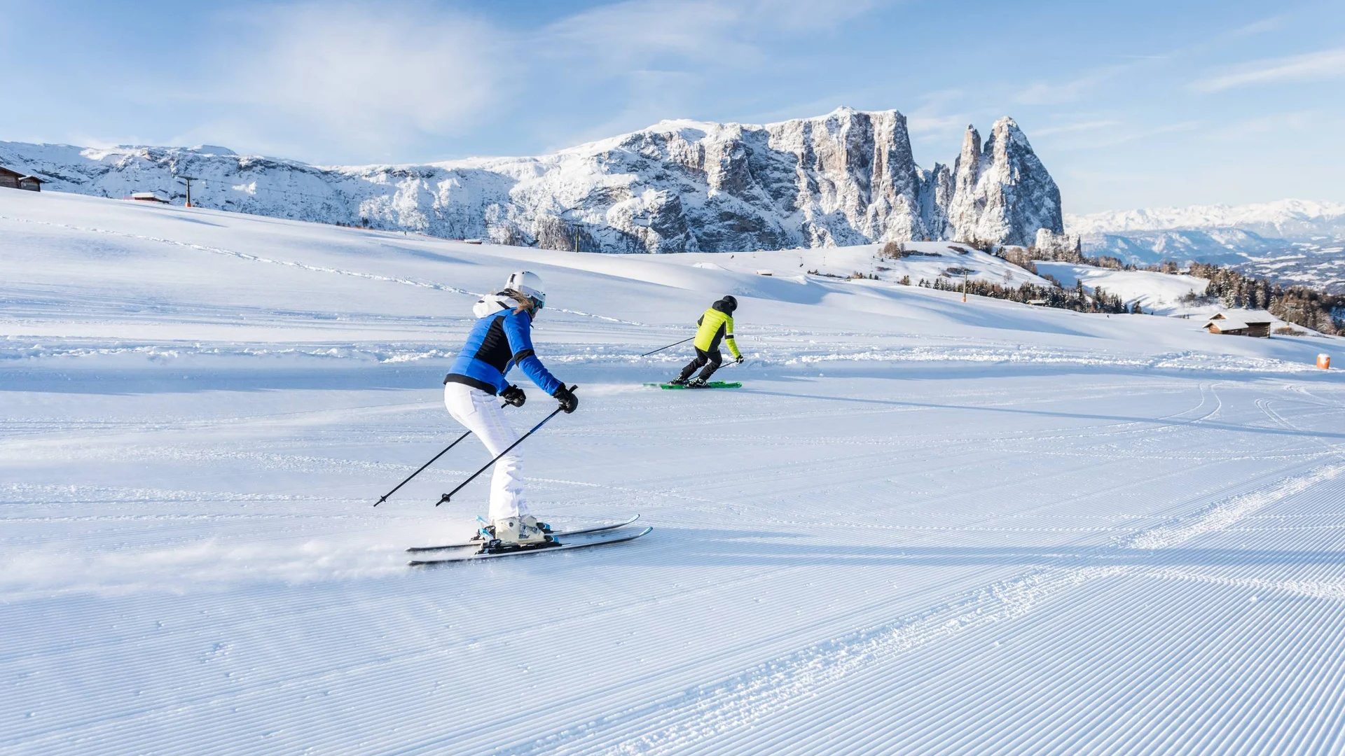 Zwei Skifahrer fahren auf einer präparierten Skipiste mit schneebedeckten Bergen im Hintergrund