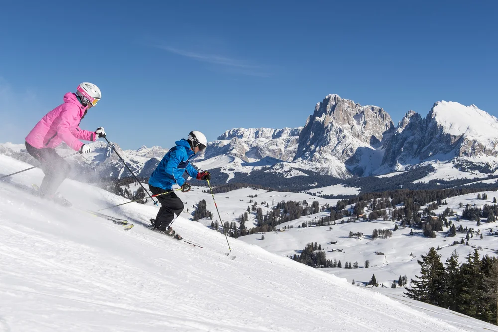 Zwei Skifahrer in bunter Kleidung fahren einen schneebedeckten Hang mit Bergkulisse hinab