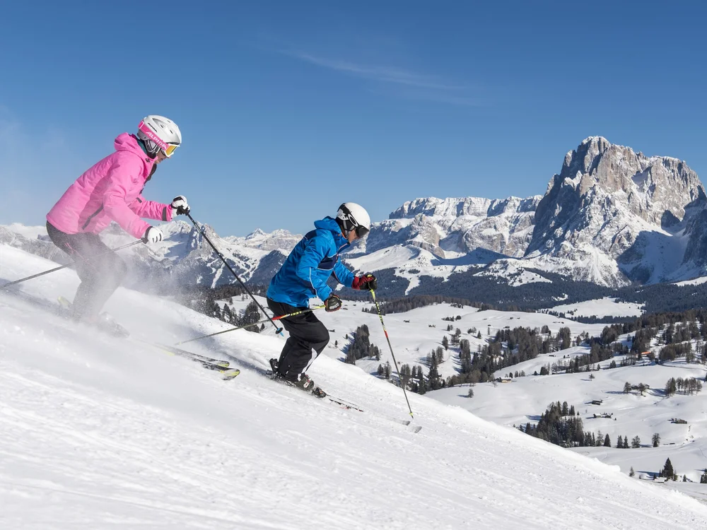 Zwei Skifahrer in bunter Kleidung fahren einen schneebedeckten Hang mit Bergkulisse hinab