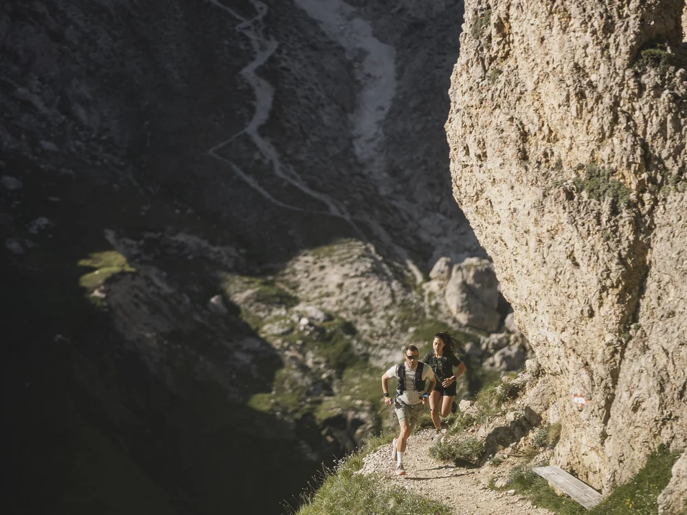 Zwei Personen laufen auf einem schmalen Bergpfad neben steiler Felswand