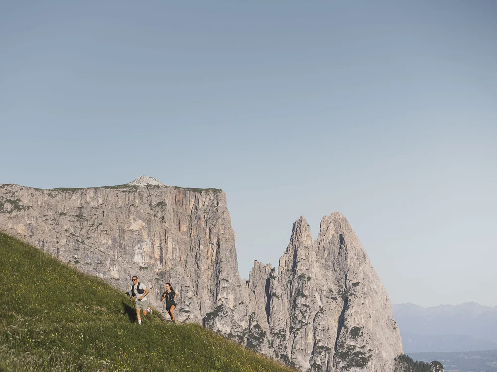 Zwei Personen laufen auf einer Wiese mit Felsen und Bergen im Hintergrund