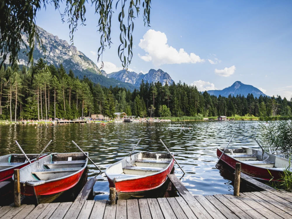 Ruderboote am Steg eines Bergsees mit Wald und Bergen im Hintergrund