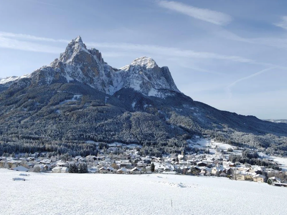 Schneebedecktes Dorf vor bewaldetem Berg unter blauem Himmel