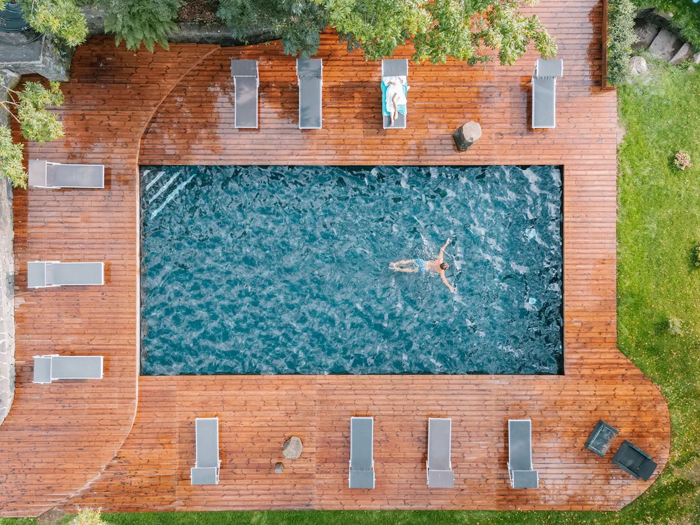 Person schwimmt in Pool neben Holzboden mit Liegestühlen und Grünfläche