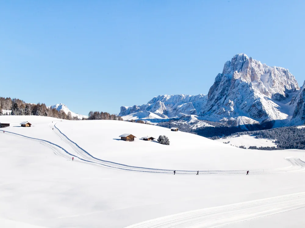 Schneebedeckte Landschaft mit Skifahrern und Berghütten vor Bergkulisse