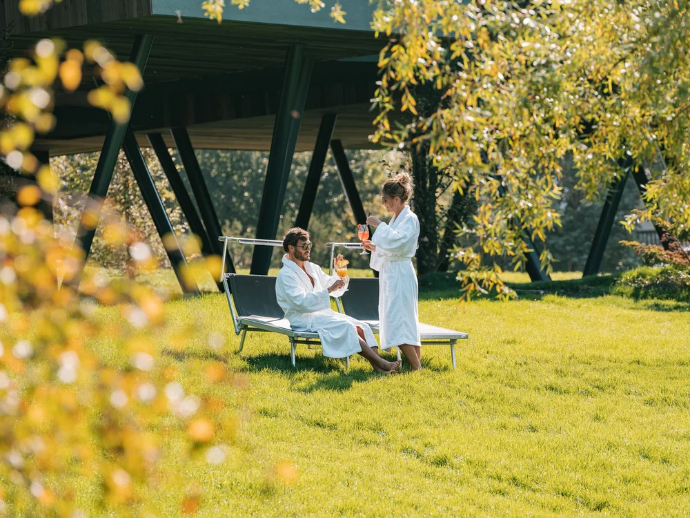 Paar in Bademänteln mit Getränken auf Liegestühlen im Garten an einem sonnigen Tag